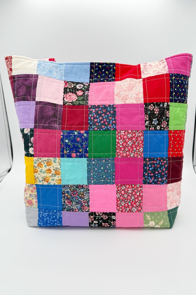 May include: A patchwork tote bag with a variety of colorful fabric squares. The squares feature floral, solid, and patterned designs in shades of pink, blue, red, green, and purple. The bag has a rectangular shape.