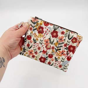 May include: A floral print pouch with a zipper closure and a silver-colored clasp. The pouch features a cream-colored background with a pattern of red, yellow, and pink flowers and green leaves. The zipper is brown.