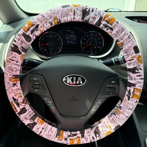 May include: A light pink steering wheel cover featuring a repeating pattern of black and orange illustrations including bats, spiders, sunflowers, potion bottles, and candles, installed on a black Kia steering wheel with a central silver logo.