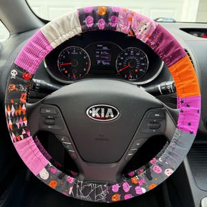 Op de afbeelding: Een stoffen stuurwielhoes met een patchworkontwerp met diverse Halloween-thema's zoals pompoenen, schedels, spoken en spinnenwebben in de kleuren oranje, paars, zwart, grijs en wit, gemonteerd op een zwart Kia-stuurwiel.