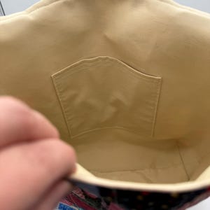 May include: Inside view of a patchwork tote bag. The interior is a light tan fabric with a small pocket. The exterior features a colorful patchwork design with various fabrics in shades of pink, blue, and black.