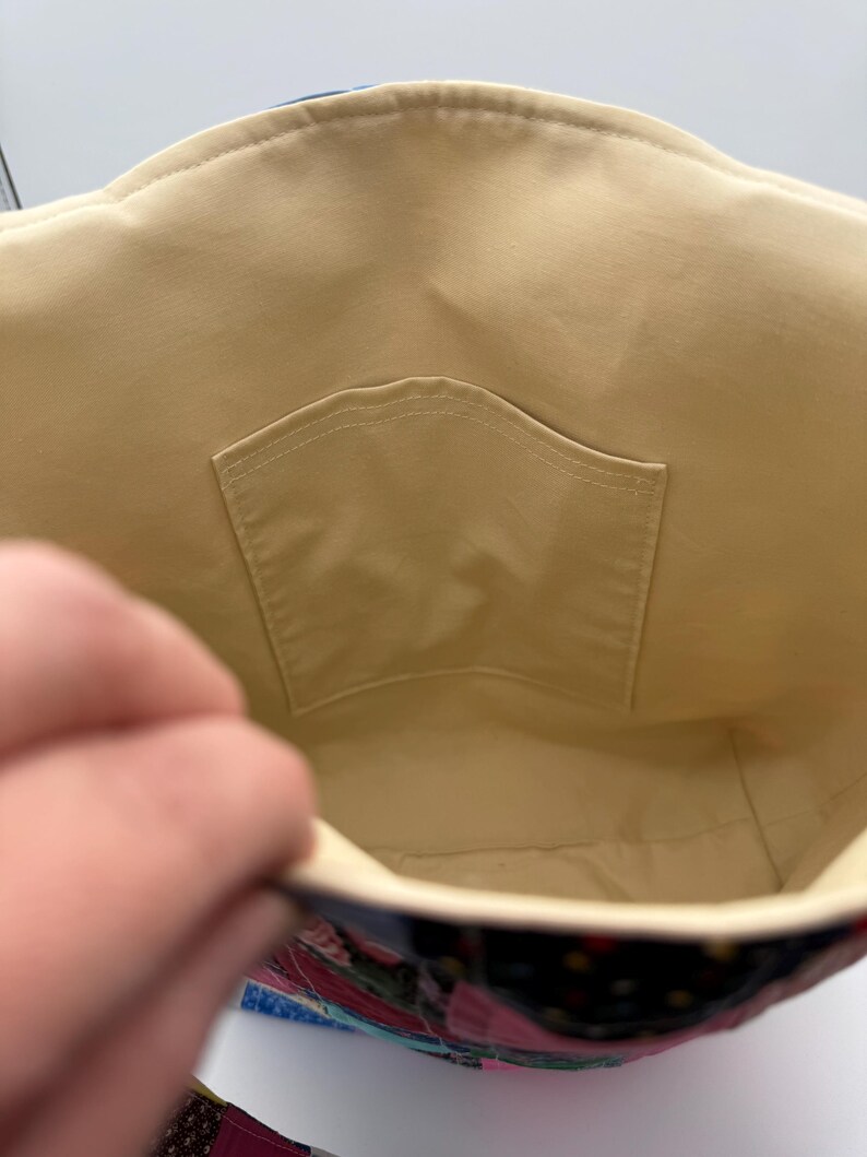 May include: Inside view of a patchwork tote bag. The interior is a light tan fabric with a small pocket. The exterior features a colorful patchwork design with various fabrics in shades of pink, blue, and black.