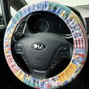 May include: A colorful patchwork steering wheel cover. The cover features a variety of fabric patterns, including floral, striped, and gingham designs in shades of blue, red, yellow, and green. The cover is designed to fit a standard car steering wheel.