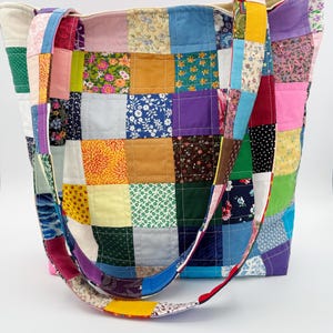 May include: A patchwork tote bag with a variety of colorful fabric squares. The bag features a mix of floral, solid, and patterned fabrics in shades of blue, green, yellow, pink, and purple. The straps are also made of patchwork fabric.