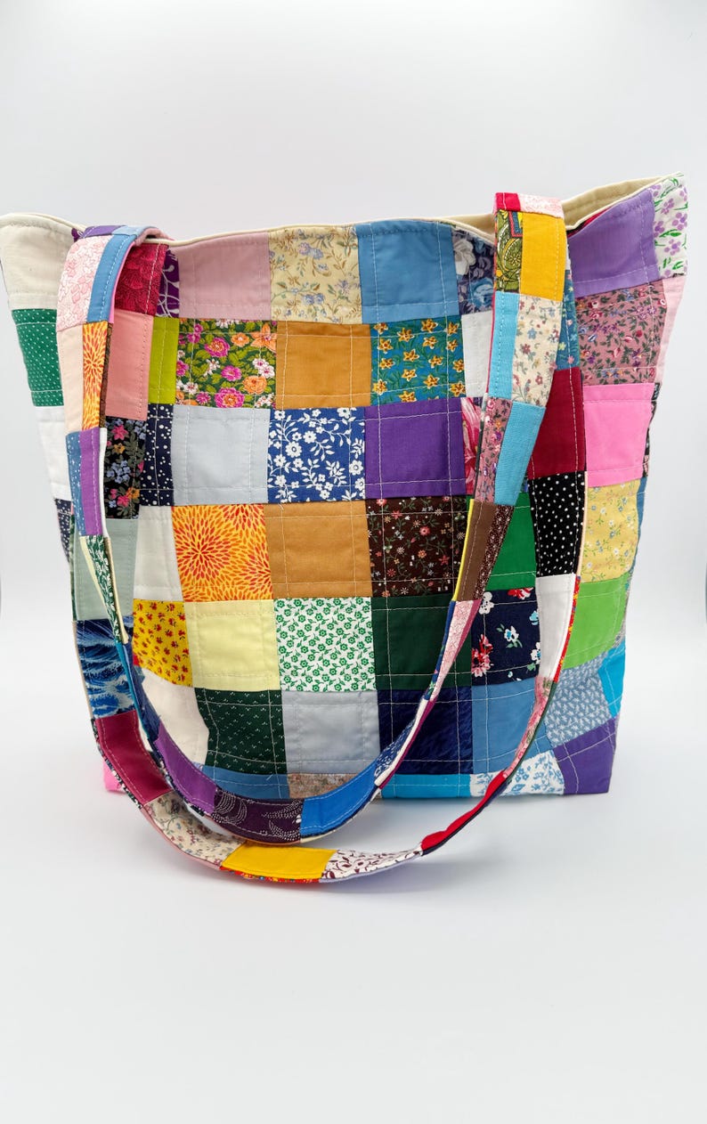 May include: A patchwork tote bag with a variety of colorful fabric squares. The bag features a mix of floral, solid, and patterned fabrics in shades of blue, green, yellow, pink, and purple. The straps are also made of patchwork fabric.