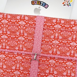 May include: A quilted laptop sleeve featuring a vibrant orange and pink pattern of moths, flowers, and foliage. The sleeve has a vertical pink floral strap with a silver metal lobster clasp closure. A silver laptop is partially visible inside the sleeve.