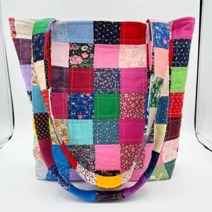 May include: A patchwork tote bag with a variety of colorful fabric squares. The bag features a rainbow-colored handle and is made from various floral and solid fabrics. The bag is designed for carrying items and has a rectangular shape.
