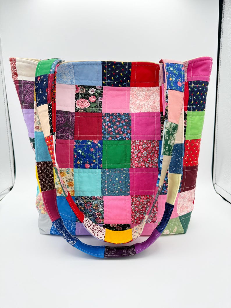 May include: A patchwork tote bag with a variety of colorful fabric squares. The bag features a rainbow-colored handle and is made from various floral and solid fabrics. The bag is designed for carrying items and has a rectangular shape.