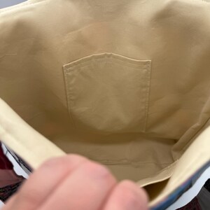May include: Interior view of a beige tote bag with a small pocket. The bag's lining is a solid beige color, and the stitching is visible along the seams. The bag's exterior features a patchwork design with various colors and patterns.