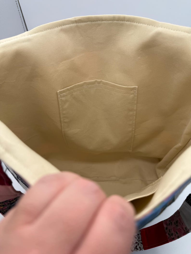 May include: Interior view of a beige tote bag with a small pocket. The bag's lining is a solid beige color, and the stitching is visible along the seams. The bag's exterior features a patchwork design with various colors and patterns.