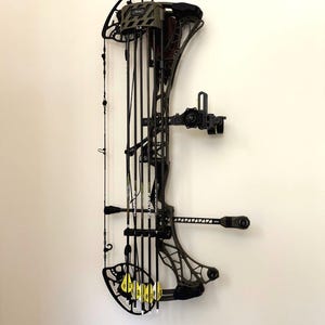 May include: A compound bow with arrows attached, mounted on a white wall. The bow is primarily black with some olive green accents. The arrows have yellow fletching. The bow is designed for target shooting or hunting.
