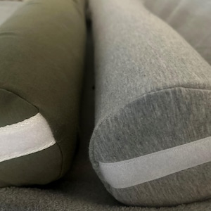 May include: Two cylindrical pillows, one green and one grey, with white trim. Both pillows have a textured fabric surface.