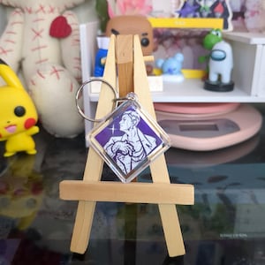May include: A purple and white square keychain with a picture of a person with a crown on their head. The keychain is on a small wooden easel.