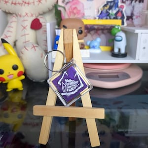 Puede incluir: Un llavero cuadrado morado y blanco con un diseño gráfico de un personaje de un videojuego. El llavero está en un pequeño caballete de madera.