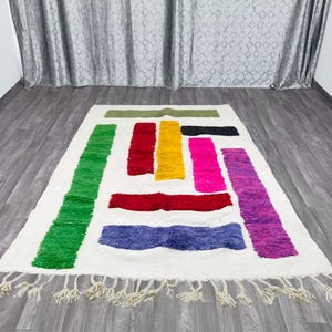 May include: A white rug with colorful geometric shapes in shades of green, red, yellow, pink, and purple. The rug has a fringe border.