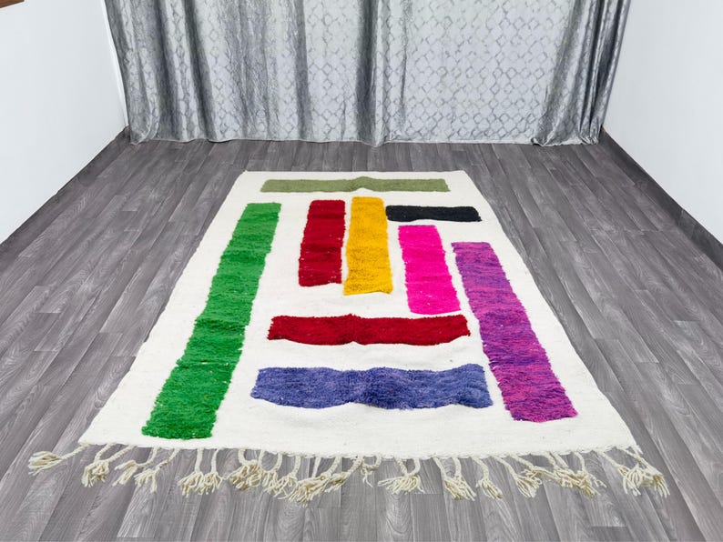 May include: A white rug with colorful geometric shapes in shades of green, red, yellow, pink, and purple. The rug has a fringe border.