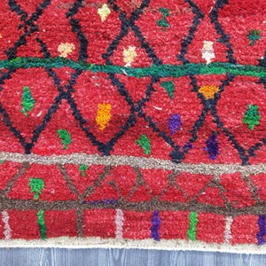 May include: Close-up of a red rug with a geometric pattern. The rug features a diamond design in black, with accents of green, yellow, and purple. The rug has a textured appearance, with a border of brown and white.