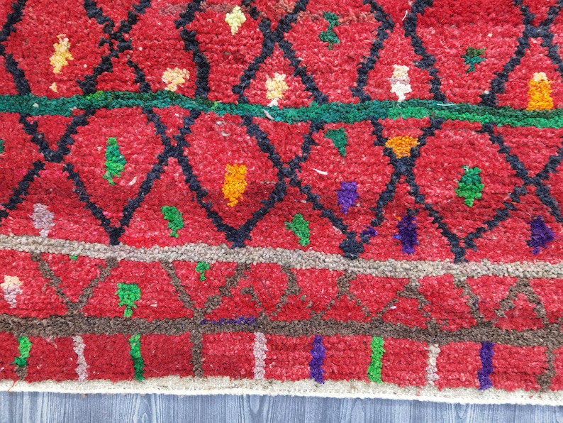May include: Close-up of a red rug with a geometric pattern. The rug features a diamond design in black, with accents of green, yellow, and purple. The rug has a textured appearance, with a border of brown and white.
