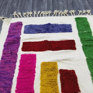 May include: A white rug with colorful stripes of purple, pink, red, yellow, and green. The rug has a fringe border.