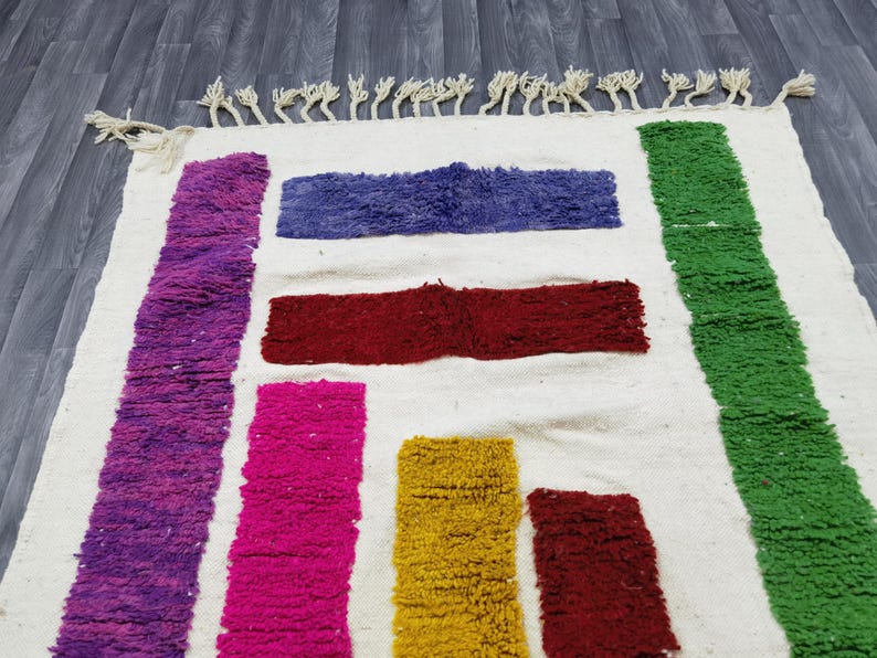 May include: A white rug with colorful stripes of purple, pink, red, yellow, and green. The rug has a fringe border.