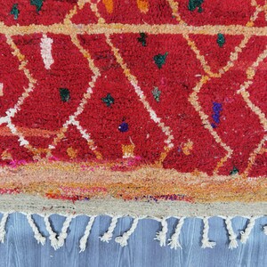 May include: Close-up of a red rug with a geometric diamond pattern in white and yellow. The rug has a textured appearance with small colorful accents and a fringe of white tassels along the bottom edge. The rug is a traditional style.