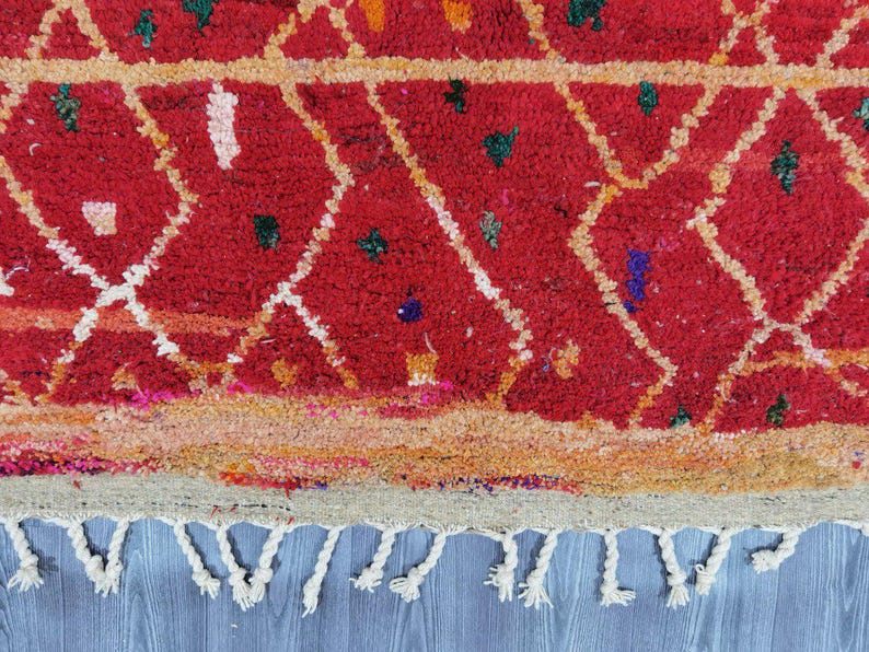 May include: Close-up of a red rug with a geometric diamond pattern in white and yellow. The rug has a textured appearance with small colorful accents and a fringe of white tassels along the bottom edge. The rug is a traditional style.