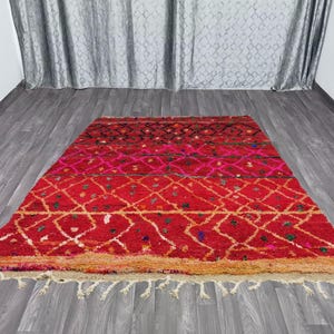 May include: A rectangular, hand-woven rug with a vibrant red and pink color scheme. The rug features a geometric diamond pattern in white and gold, with a fringe along the bottom edge. The rug is made of wool and is approximately 5 feet by 7 feet.