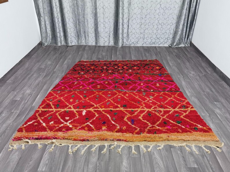 May include: A rectangular, hand-woven rug with a vibrant red and pink color scheme. The rug features a geometric diamond pattern in white and gold, with a fringe along the bottom edge. The rug is made of wool and is approximately 5 feet by 7 feet.