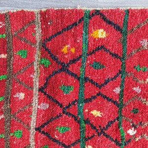 May include: Close-up of a red, textured rug with a geometric pattern. The design features black diamond shapes, green vertical lines, and accents of yellow, green, and white. The rug has a thick, woven texture.