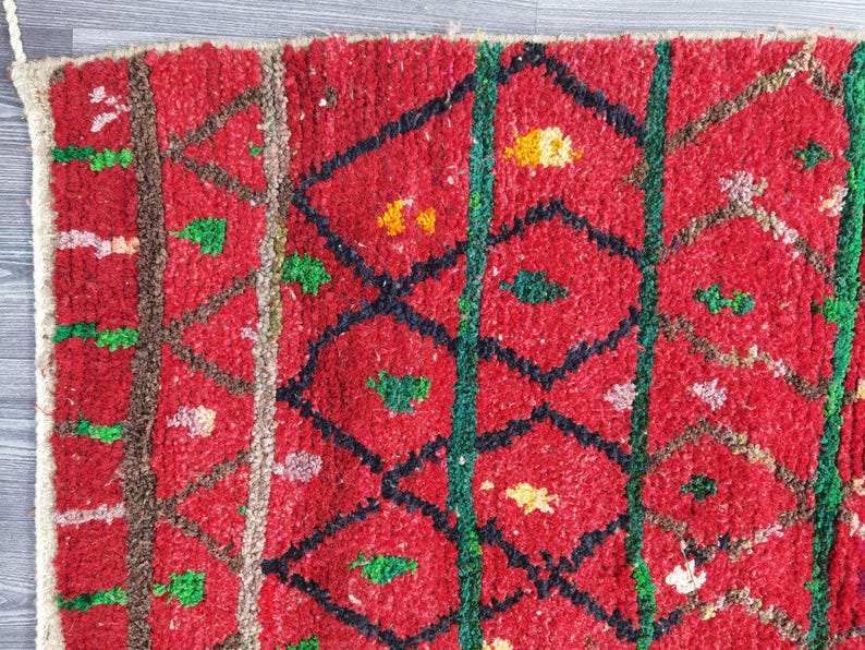 May include: Close-up of a red, textured rug with a geometric pattern. The design features black diamond shapes, green vertical lines, and accents of yellow, green, and white. The rug has a thick, woven texture.