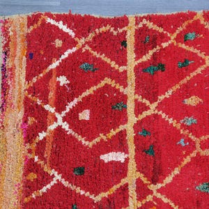 May include: Close-up of a vibrant red rug with a geometric diamond pattern. The rug features white and yellow lines, along with small green and orange accents. The edge has a beige fringe.