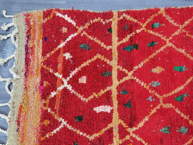 May include: Close-up of a vibrant red rug with a geometric diamond pattern. The rug features white and yellow lines, along with small green and orange accents. The edge has a beige fringe.