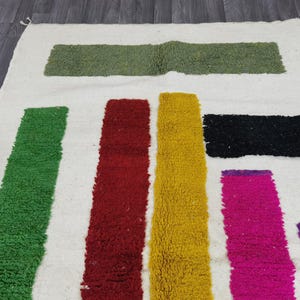 May include: A white rug with colorful stripes of green, red, yellow, pink, and purple. The stripes are woven into the rug and create a geometric pattern.