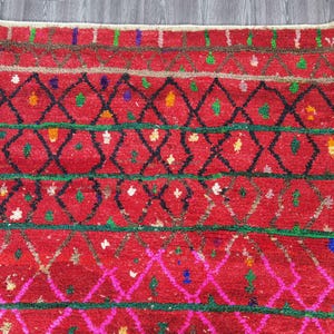 May include: A vibrant red rug with a geometric pattern. The rug features black diamond shapes, green and pink lines, and colorful accents. The rug's design is intricate and eye-catching, suitable for adding a pop of color and texture to any room.