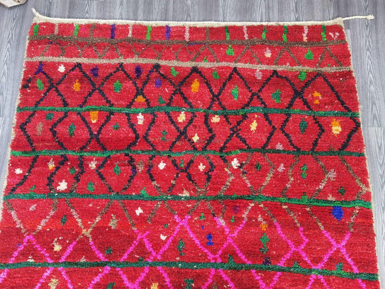 May include: A vibrant red rug with a geometric pattern. The rug features black diamond shapes, green and pink lines, and colorful accents. The rug's design is intricate and eye-catching, suitable for adding a pop of color and texture to any room.