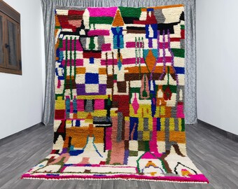 May include: A large, colorful rug with an abstract geometric design. The rug features a variety of shapes and colors, including pink, green, yellow, and blue. The rug is made of a thick, textured material and is displayed in a room with gray flooring.