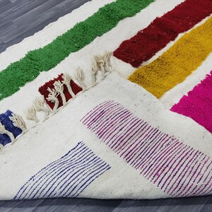 May include: A white rug with colorful stripes of green, red, yellow, pink, and purple. The rug has a fringe border and is made of a soft, fluffy material.