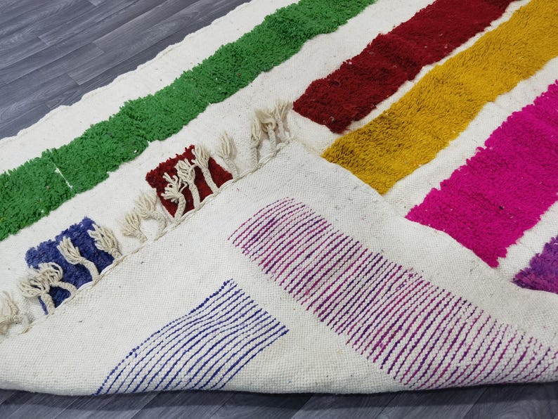May include: A white rug with colorful stripes of green, red, yellow, pink, and purple. The rug has a fringe border and is made of a soft, fluffy material.