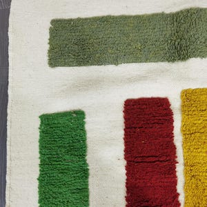 May include: A white rug with green, red, and yellow stripes. The stripes are woven into the rug and create a geometric pattern.