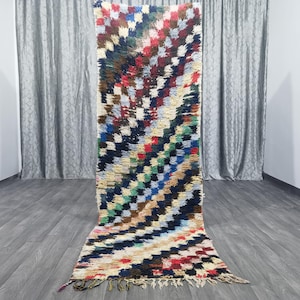 May include: A colorful Moroccan rug with a geometric pattern in shades of red, blue, green, and white. The rug has a fringe border and is made of wool.