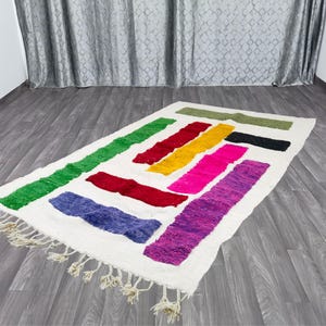 May include: A white rug with colorful stripes in shades of green, yellow, pink, purple, and red. The rug has fringe around the edges.