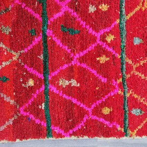 May include: Close-up of a vibrant red rug with a geometric pattern. The design features bright pink diamond shapes, green vertical lines, and small accents of gold and other colors. The rug has a textured, woven appearance.