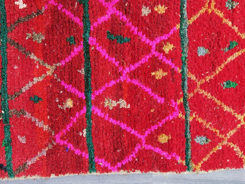 May include: Close-up of a vibrant red rug with a geometric pattern. The design features bright pink diamond shapes, green vertical lines, and small accents of gold and other colors. The rug has a textured, woven appearance.