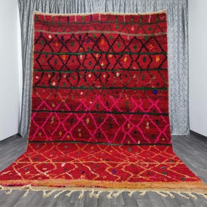 May include: A vibrant red rug with geometric diamond patterns in black, pink, and green. The rug has a textured surface and a fringed edge. The design features a gradient from a lighter orange at the bottom to a darker red at the top. This rug is suitable for home decor.