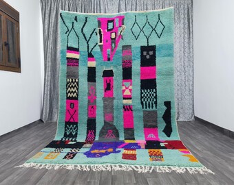 May include: A turquoise, rectangular rug with a geometric design. The rug features vertical columns in black, pink, gray, and white, with various patterns. The bottom edge has a fringe. The rug is displayed indoors.