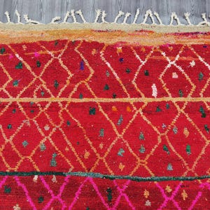 May include: A vibrant red rug with a geometric diamond pattern in white and gold. The rug features a textured surface with small colorful accents and a fringed edge. The design includes a pink and green border.