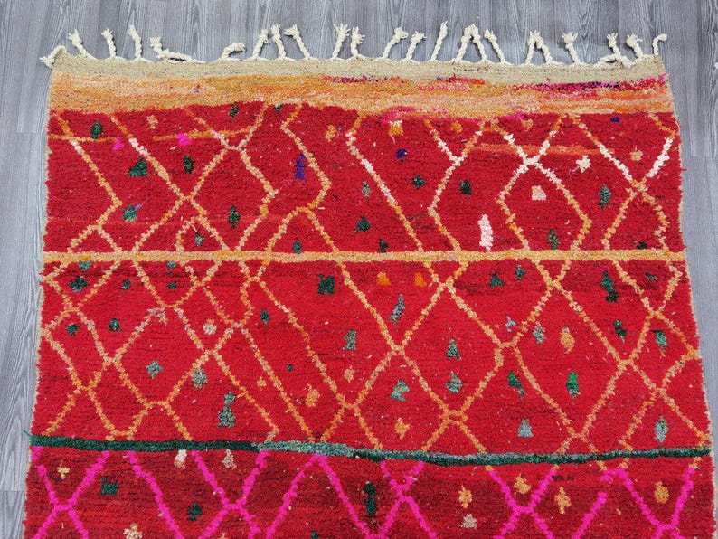 May include: A vibrant red rug with a geometric diamond pattern in white and gold. The rug features a textured surface with small colorful accents and a fringed edge. The design includes a pink and green border.