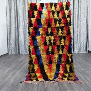 May include: A colorful Moroccan rug with a geometric pattern in shades of red, yellow, blue, and black. The rug has a fringe border and is made of wool.