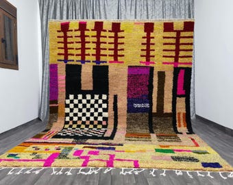 May include: A large, colorful rug with a geometric design. The rug features a yellow background with rectangular shapes in black, brown, pink, and purple. A checkered pattern is also present. The rug has a fringe along the bottom edge.