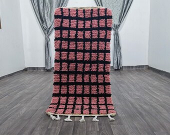 Könnte beinhalten: Ein rechteckiger Teppich mit einem rosa-schwarzen Karomuster. Der Teppich hat eine zottelige Textur und ist unten mit weißen Quasten verziert. Das Design zeigt ein Gitter aus schwarzen Linien auf rosa Hintergrund.